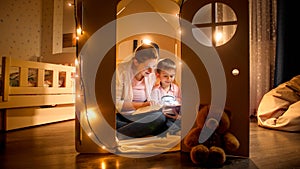 Young smiling woman reading story book to her little son at night while sitting on floor. Concept of child education and