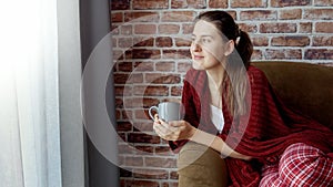 Young smiling woman relaxing on sofa with cup of tea at big window. Female resting at home, lifestyle and relaxation