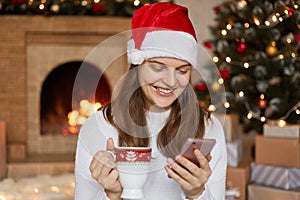 Young smiling woman in santa claus hat smiling and sending greetings sms using smart phone on fireplace and christmas tree
