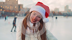 Young smiling woman Santa hat ice skating outside on ice rink dressed white sweater. Christmas holiday, active winter