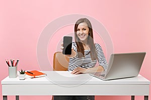 Young smiling woman showing on camera mobile phone with blank empty screen work at white desk with contemporary pc