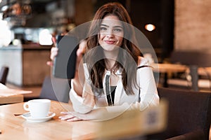 Young smiling woman showing her smartphone during lunch break drinking hot coffee in trendy cafe looking happy modern