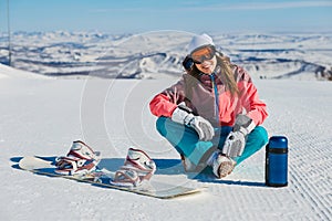 A young smiling woman sits on a mountain slope with a snowboard and a thermos