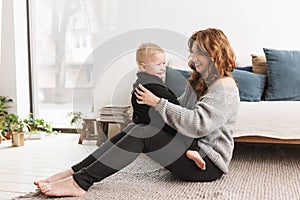 Young smiling woman sitting on floor happily playing with her little handsome son. Mom with baby boy joyfully spending