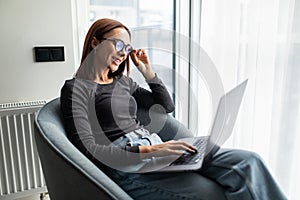 Young smiling woman sitting on modern chair near the window in light cozy room at home working on laptop in relaxing atmosphere