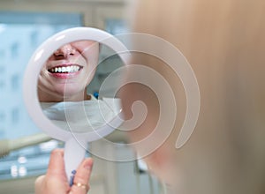 Young smiling woman sitting in stomatology clinic chair and looked at mirror evaluating her teeth reflection after Tooth whitening