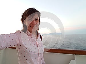 Young smiling woman in soft pink shirt making selfie photo on board of cruise ship at open deck to share on internet