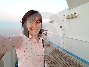 Young smiling woman in soft pink shirt making selfie photo on board of cruise ship at open deck to share on internet social media