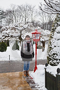 Young smiling woman standing near an old style red lantern with christmas decoration in it in a snow-covered winter park. Snowy