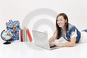 Young smiling woman student in denim clothes working on laptop pc computer lying near globe backpack and school books