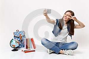 Young smiling woman student doing taking selfie shot on mobile phone showing victory sign near globe, backpack, school