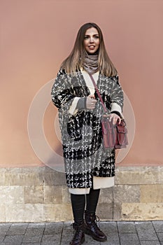 Young smiling woman in a stylish coat and with a red bag on the city street. Beautiful blonde on a background of a pink wall.