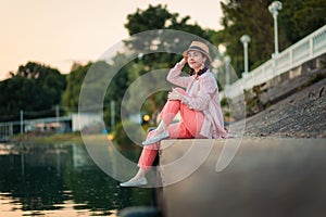 A young smiling woman in summer clothes and straw hat sitting at river embankment. Summer vacation and relax
