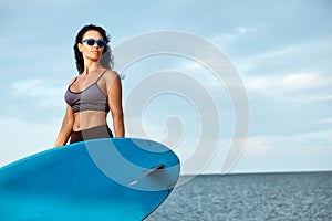 Young smiling woman in sunglasses with surfboard standing on the beach