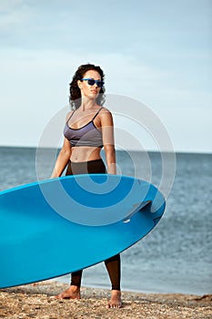 Young smiling woman in sunglasses with surfboard standing on the beach