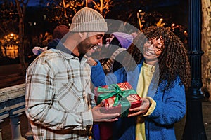 Young smiling woman surprising and giving a christmas gift to her boyfriend on a happy party with friends. Pleased man