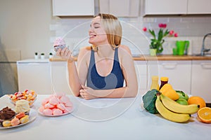 Young smiling woman with sweet cake in one hand choosing between healthy and unhealthy food in the kitchen. Difficult