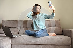 Young smiling woman taking a selfie for her profile. She sitting on  couch close to laptop at home. lockdown and online dating con