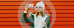 Young smiling woman taking selfie picture by phone wearing a knitted hat, sweater on red