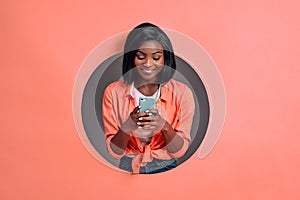 Young smiling woman typing text message or using internet on smartphone isolated in a round hole in coral background. Copy space