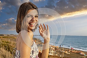 Young smiling woman waving at the spectator. Cheerful gesture. Seaside landscape in the background.