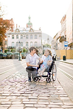 Young smiling woman in wheelchair and joyful man walking outdoors in old city center in summer morning