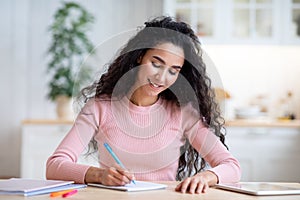 Young Smiling Woman Writing Notes In Notepad, Sitting At Table In Kitchen