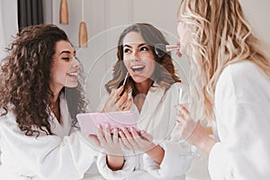Young smiling women 20s wearing housecoat sitting in luxury bedroom at hotel room, and applying makeup for bride during bridal sh photo