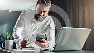 Young smilling bearded businessman standing near table in front of laptop, using smartphone. Man checks e-mail