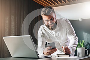 Young smilling bearded businessman standing near table in front of laptop, using smartphone. Man checks e-mail