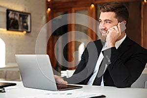Young smliing bearded business man in black suit talking on mobile phone, looking aside in office