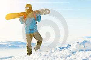 Young snowboarder in helmet standing at the top of a mountain with sun behind him