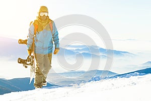 Mladý snowboardista so slnkom za ním kráčajúci do hory drží v ruke snowboard