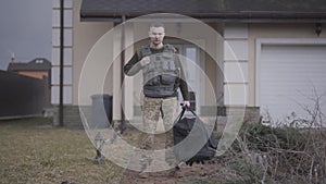 The young soldier in military closes with big bag standing in front of the house. A man going to the military service