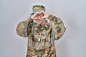 Young south asian woman wearing camouflage army uniform doing heart shape with hand and fingers smiling looking through sign