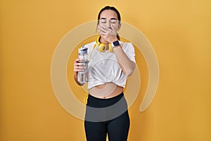 Young south asian woman wearing sportswear drinking water bored yawning tired covering mouth with hand