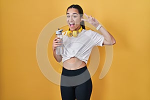 Young south asian woman wearing sportswear drinking water doing peace symbol with fingers over face, smiling cheerful showing