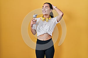 Young south asian woman wearing sportswear drinking water smiling confident touching hair with hand up gesture, posing attractive