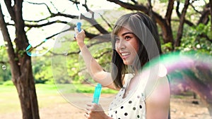 Young South East Asian ethnic 20s woman blowing air bubbles in the park. Happy girl wears summer dress with trees in