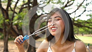 Young South East Asian ethnic 20s woman blowing air bubbles in the park. Happy girl wears summer dress with trees in