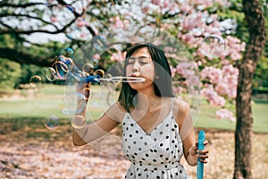 Young South East Asian ethnic 20s woman blowing air bubbles in the park. Happy girl wears summer dress with trees in