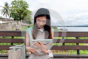Young Southeast Asian woman using a tablet computer while sitting on a park bench