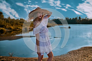 Young spectacular blonde in a straw hat and white dress posing on the river bank in summer in clear sunny weather