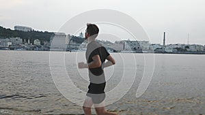 Young sportive boy in black sports apparel running along river beach. Active slim man runs near river. Athlete doing outdoor exerc
