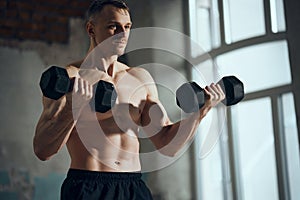 Young sportive man with strong muscular, relief, body training shirtless, lifting dumbbells indoors on daytime. Concept