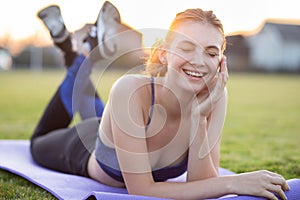 Young sportive woman in sports clothes laying on training mat before doing exercises in field at sunrise