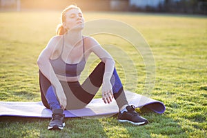 Young sportive woman in sports clothes sitting on training mat before doing exercises in field at sunrise