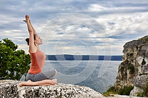 Young sportive woman training yoga asanas on rock above beautiful river.