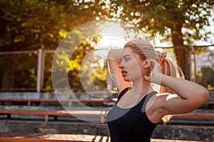 Young sportive woman tying her hair into pony tail on sportsground in summer. Preparation for workout