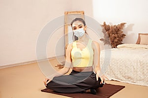 Young sports girl in beige interior. Yellow grey clothing. A healthy lifestyle. A woman in a medical mask is engaged in yoga